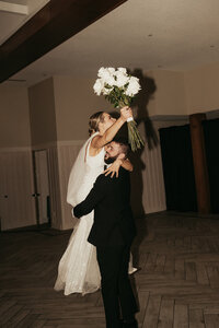 Groom lifts bride in the air as bride raises her bouquet in a celebratory way