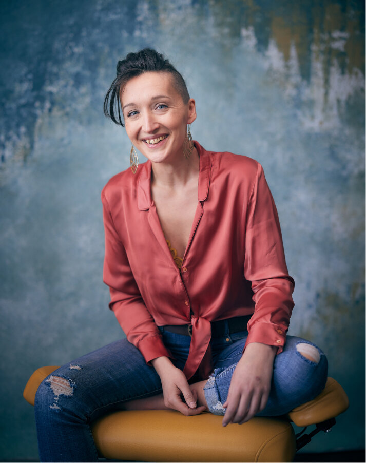 Tarzan, a slim, 38-year-old white woman in a peach low-collared silk dress shirt and ripped jeans, sits on a yellow office chair and smiles meaningfully